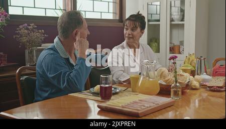 Una coppia di mezza età che chiacchiera durante la colazione. Sono seduti al sontuoso tavolo da cucina, sorridendo e indossando pigiama. Coppia sposata in conversin Foto Stock