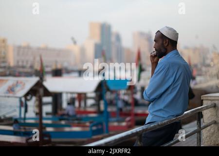 Un barbuto con Kufi si appoggia su una ringhiera con una sigaretta tra le labbra. L'operatore Abra fuma durante la sua pausa. I taxi acquei attraccati sullo sfondo. Foto Stock