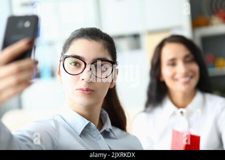 La giovane donna in occhiali tiene il telefono davanti al viso e sorride ai colleghi in ufficio Foto Stock