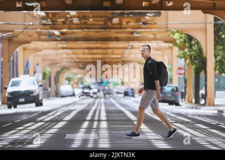 Vista laterale di un adulto con zaino a piedi su una strada pedonale a crosswalk e su una ferrovia sopraelevata dei mezzi pubblici. Chicago, Stati Uniti Foto Stock