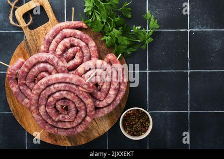 Salsicce di maiale crude a spirale. Salsicce di maiale fresco gustoso spirale ritorta per barbecue su stativo bianco con spezie ed erbe per la cena di festa Oktoberfest sul b grigio Foto Stock