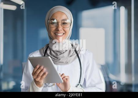 Ritratto di una dottoressa musulmana in un hijab con una tavoletta in mano, il medico lavora in ufficio di una clinica moderna, sorride e guarda la macchina fotografica. Foto Stock