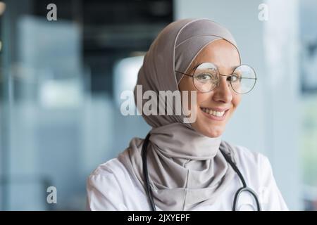 Primo piano ritratto fotografico di una dottoressa musulmana in hijab grigio, medico femminile sorridente e guardando la macchina fotografica, operatrice in occhiali e camice medico bianco e occhiali che lavorano all'interno di un moderno ufficio clinico Foto Stock