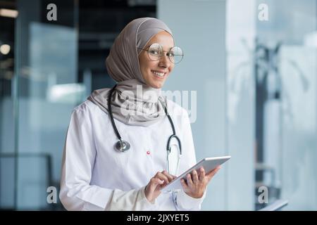 Ritratto di una dottoressa musulmana in un hijab con una tavoletta in mano, il medico lavora in ufficio di una clinica moderna, sorride e guarda la macchina fotografica. Foto Stock