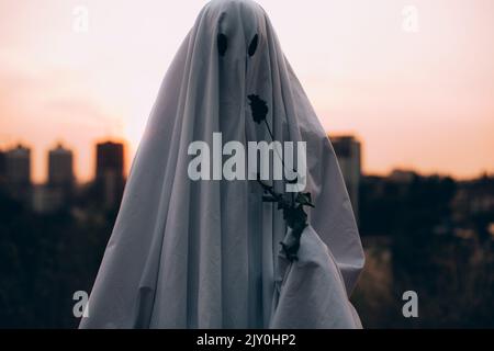 Fantasma che tiene il fiore selvaggio in piedi contro la città di sera durante Halloween Foto Stock