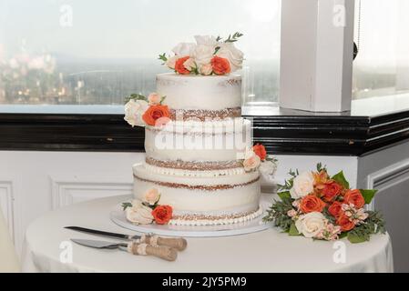 La torta nuziale a tre strati è situata accanto alla finestra con vista sulla città, insieme a un set di servizio e un bouquet di fiori. Foto Stock