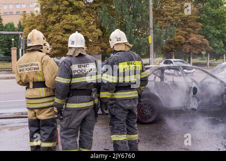 Salvatore in uniforme con l'iscrizione del Ministero degli Affari interni della Russia in russo. I vigili del fuoco sono in piedi accanto all'auto bruciata. Foto Stock