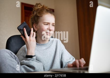 Ragazza caucasica dai capelli rossi bella adolescente in una felpa con cappuccio grigia e pantaloni, seduta a un tavolo di fronte a un computer portatile con uno smartphone. Una ragazza con divertente Foto Stock