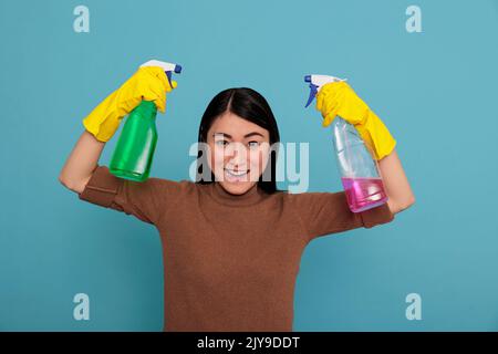 Eccitata glade e ottimista governante asiatico al lavoro che solleva le braccia tenendo due spray detergenti nei guanti gialli, pulizia concetto casa, allegra compiaciuta e positiva casalinga da chores Foto Stock