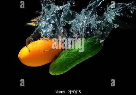 Cetriolo verde fresco e pomodoro giallo caduto in acqua con spruzzi isolati su sfondo nero. Foto Stock