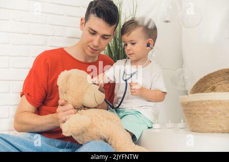 Giochi diversi con i bambini. Primo piano medio sparato al coperto di papà millenario caucasico con il suo figlio preschooler che gioca medico con orsacchiotto. Stetoscopio. Foto di alta qualità Foto Stock