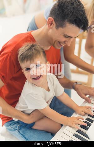 Concetto di infanzia divertente. La musica nella vita dei bambini. Felice sorridente orgoglioso papà caucasico dai capelli scuri che suona il pianoforte con il suo sorridente al figlio della macchina fotografica. Rapporto padre-figlio. Foto di alta qualità Foto Stock