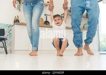 Basso colpo indoor di due adulti caucasici sposati in jeans blu chiaro tenendo le mani del loro figlio carino dai capelli castani vestito con t-shirt bianca. Passeggiata a piedi nudi in cucina. Foto di alta qualità Foto Stock