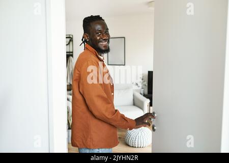 Benvenuto a casa mia. Allegro uomo nero che cammina nel suo appartamento, entrando in una nuova casa e guardando indietro la macchina fotografica Foto Stock