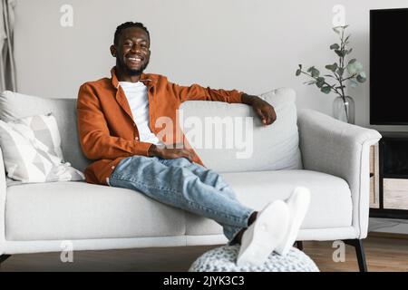 Rimani a casa da solo, goditi il silenzio. Felice uomo afroamericano seduto sul divano in un accogliente soggiorno luminoso, spazio copia Foto Stock
