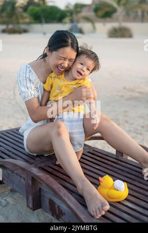 Bambino in vacanza al mare seduto sul lettino con la madre al tramonto. Donna asiatica e il suo bambino di un anno che si divertono in vacanza estiva Foto Stock