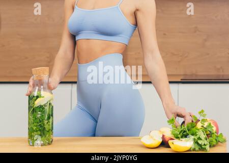 Primo piano foto parte del corpo della giovane donna fitness in piedi e tenere una bottiglia con succo fresco, disintossicazione e verdure fresche a casa in cucina Foto Stock