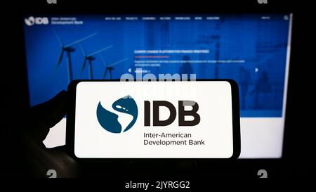 Persona che tiene in mano uno smartphone con il logo della Inter-American Development Bank (IADB) sullo schermo di fronte al sito Web. Messa a fuoco sul display del telefono. Foto Stock