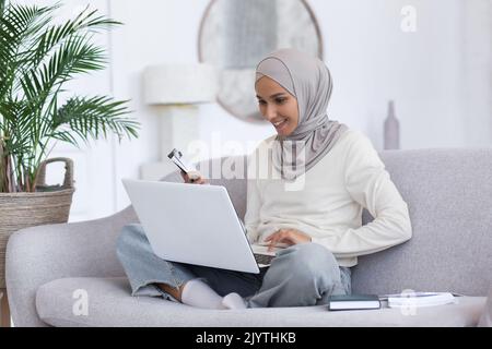 Giovane bella donna musulmana in hijab grigio studiando a casa seduta sul divano, studente femmina che utilizza il computer portatile per l'apprendimento a distanza, guardando la lezione in salotto. Foto Stock