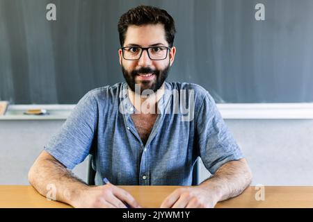 Allegro ritratto dell'insegnante maschile seduto alla scrivania in aula Foto Stock