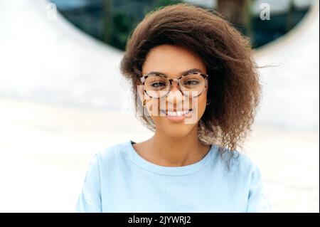 Primo piano di una bella donna afro-americana positiva e sicura con capelli ricci, indossata in una camicia blu, con occhiali, in piedi all'aperto, guardando la macchina fotografica, sorridente amichevole Foto Stock