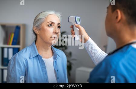 Medico asiatico adulto maschio controllo della temperatura con termometro senza contatto a paziente anziano triste signora Foto Stock