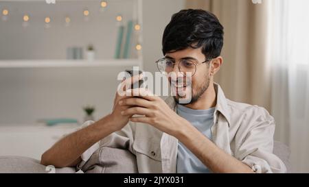 Giovane uomo 30s arabo barbuto uomo millenario in occhiali si siede a casa sul divano guardando il telefono cellulare facendo sì gesto ottenere offerta vincendo buone notizie Foto Stock