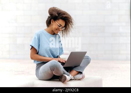 Utilizzo di laptop, tecnologia wireless. Donna afroamericana intelligente, freelancer femminile di razza mista, studente, con occhiali, in camicia blu, lavorando in un computer portatile mentre si siede all'aperto, guarda lo schermo, sorridere Foto Stock