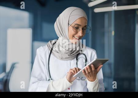 Medico femminile in hijab lavora in clinica moderna ufficio, medico musulmano donna utilizza tablet computer, infermiere in camice medico bianco e occhiali con stetoscopio. Foto Stock