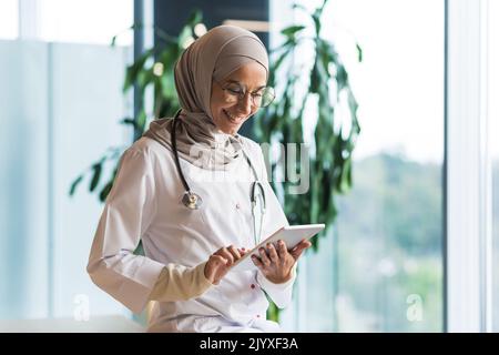 Medico femminile in hijab lavora in clinica moderna ufficio, medico musulmano donna utilizza tablet computer, infermiere in camice medico bianco e occhiali con stetoscopio. Foto Stock