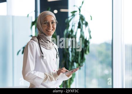 Ritratto di una dottoressa musulmana in un hijab con una tavoletta in mano, il medico lavora in ufficio di una clinica moderna, sorride e guarda la macchina fotografica. Foto Stock