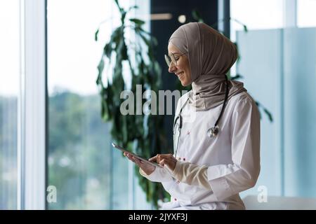 Medico femminile in hijab lavora in clinica moderna ufficio, medico musulmano donna utilizza tablet computer, infermiere in camice medico bianco e occhiali con stetoscopio. Foto Stock