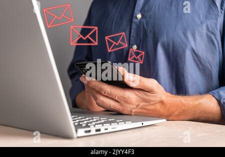 Uomo che riceve e-mail di phishing al telefono mentre lavora sul computer portatile. Maschio seduto al tavolo con il computer e tenendo lo smartphone. Messaggi indesiderati, spam te Foto Stock