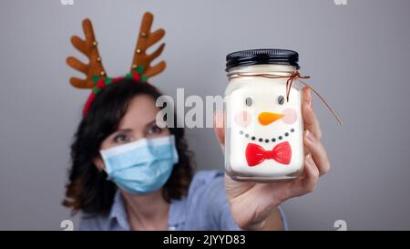 Donna con maschera e fascia di renna che mostra pupazzo di neve. Può con un volto di Natale. Maschera medica, scatto di primo piano, fuoco selettivo, prevenzione dal pandem Foto Stock