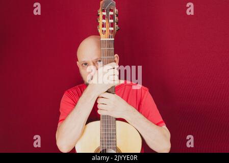 Strumento musicale a corda. Chitarrista maschile carismatico maturo. Ragazzo con barba tiene la chitarra. Uomo bearded in una camicia rossa. Musicista esecutore di musica. Foto Stock