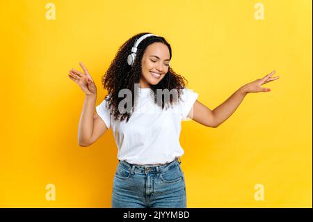 Allegro bella giovane bruna mista donna da corsa, in abbigliamento casual elegante, con cuffie, ascolta la sua musica preferita, ballare con gli occhi chiusi, rilassarsi, divertirsi su sfondo arancione isolato, sorridere Foto Stock