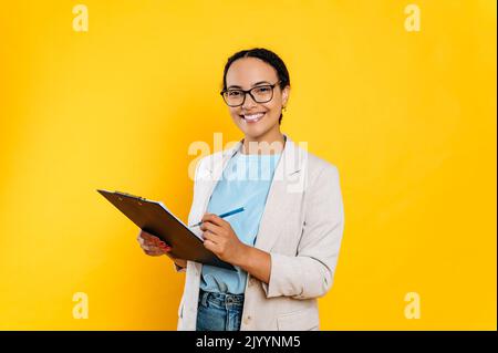 Elegante sicuro successo bella donna d'affari brasiliana o ispanica, agente immobiliare, manager finanziario, in abbigliamento formale, stand su sfondo isolato, tiene documenti, guarda la fotocamera, sorrisi Foto Stock