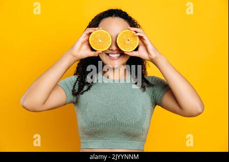 Cibo sano e stile di vita, detox. Gioiosa bella gara mista giovane donna dai capelli ricci, in abiti sportivi, in piedi su sfondo arancione isolato, tiene due metà arancione nelle sue mani vicino agli occhi, sorridendo Foto Stock