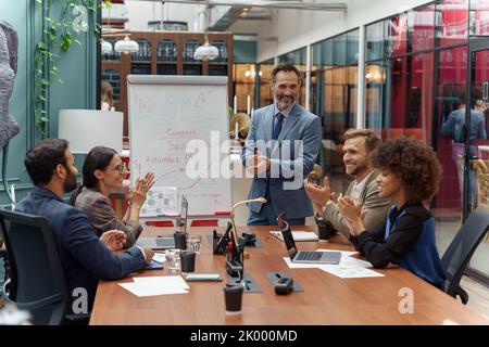 Allenatore o relatore maturo fare una presentazione su lavagna a fogli mobili per diversi uomini d'affari durante la riunione Foto Stock