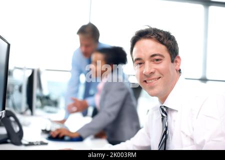 Sono orgoglioso di quello che faccio. Uomo d'affari sorridente seduto alla sua scrivania con i suoi colleghi che lavorano in background - ritratto. Foto Stock