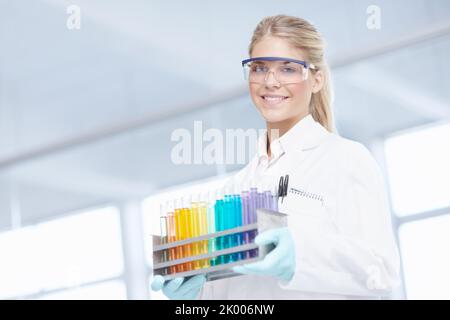 Lavoro in laboratorio. Ritratto di una bella scienziata femminile che porta un vassoio di provette mentre sorride alla macchina fotografica. Foto Stock