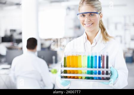 Assistente di laboratorio. Ritratto di un bel giovane scienziato che porta un vassoio di provette con un collega maschio sullo sfondo. Foto Stock