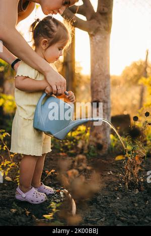 Madre aiutando la sua bambina che innaffia i fiori con annaffiatura canin giardino. Bellissimo tramonto. Assistenza ai bambini. Guardando un sacco di fiori, la famiglia Foto Stock