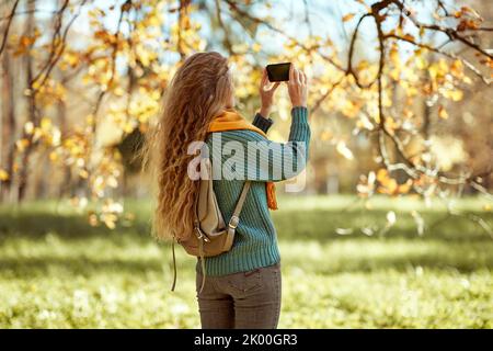 Una giovane donna dai capelli rossi cammina attraverso il parco autunnale. Una ragazza con uno smartphone scatta foto in una foresta autunnale Foto Stock