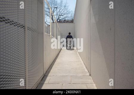 Uomo su sedia a rotelle, in avvicinamento all'edificio che si sposta lungo una rampa accessibile per persone con disabilità Foto Stock