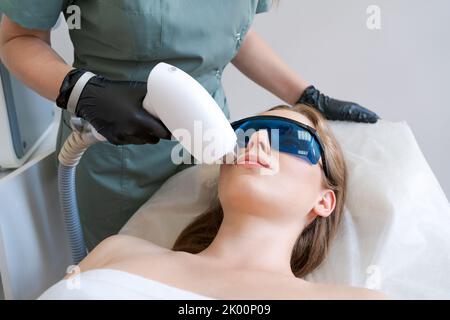 Il cosmetologo del terapista subisce il trattamento laser sulla giovane donna del viso in clinica di bellezza. Capelli laser facciali. Procedure di epilazione. Cliente soddisfatto, attenzione selettiva. Foto Stock