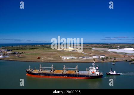 Vista aerea di una grande nave da carico guidata da un rimorchiatore vicino agli impianti portuali industriali sotto un cielo azzurro. Port Bundaberg, Queensland, Australia Foto Stock