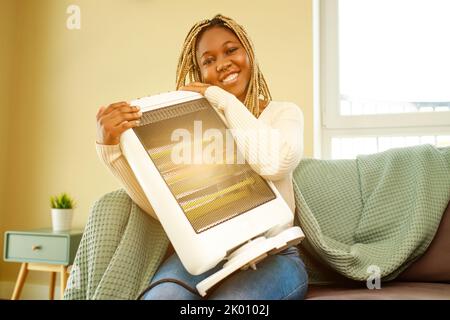 Donna afroamericana in coperta blu riscaldamento vicino al riscaldatore elettrico a casa Foto Stock