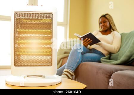 Donna afroamericana in coperta blu che legge un libro vicino al riscaldatore elettrico nel paese Foto Stock