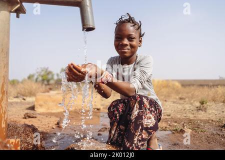 Ragazza africana assetata con trecce divertenti, seduta in mezzo a una pudle, felice di raccogliere l'acqua abbondante che scorre dalla fontana del villaggio; c Foto Stock